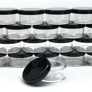 May include: A stack of clear plastic jars with black lids. The jars are arranged in a pyramid shape. There is one jar in the front that is open, showing the inside of the jar.