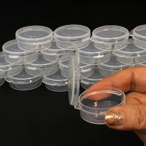 May include: A collection of small, clear plastic round containers with hinged lids. One container is open, revealing the interior. The containers are stacked, suggesting storage or organization. The background is black.