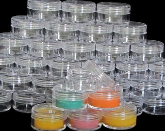 May include: A large stack of clear plastic containers with lids. The containers are arranged in a pyramid shape. There are four smaller containers in the front, filled with different colored substances.
