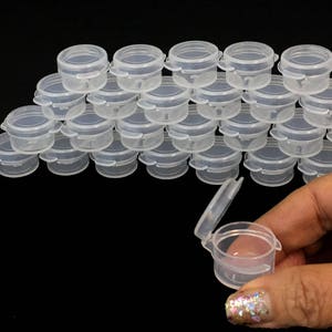 May include: A group of 24 clear plastic containers with lids. The containers are stacked in a pyramid shape. One container is open and being held by a hand.