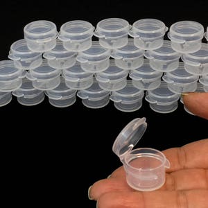 May include: A group of 20 clear plastic containers with lids. The containers are small and round, and they are arranged in a circle. One container is open and is being held by a hand.