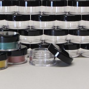 May include: A group of clear plastic jars with black lids. The jars are stacked in rows and some are filled with different colored powders.