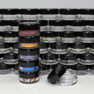 May include: A stack of small, clear, round containers with black lids. The containers are filled with various colorful items, such as glitter, sprinkles, and beads. The containers are arranged in a grid pattern on a white surface.