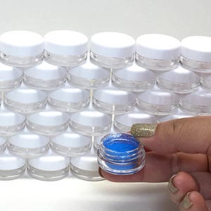 May include: A close-up of a small, clear jar filled with bright blue glitter. The jar is being held by a hand, and there are many other similar jars in the background.