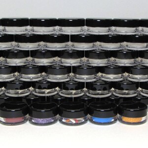 May include: A stack of small, clear plastic jars with black screw-on lids. Several jars at the bottom contain colorful contents, including pink, purple, and orange glitter, and blue and orange powder. The jars are arranged in a grid pattern.