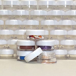 May include: A group of clear plastic containers with white lids. The containers are stacked in a pyramid shape. Some of the containers are filled with colorful sprinkles, glitter, and other small items.