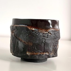 Könnte beinhalten: Ein kleiner, zylindrischer Keramikbecher mit einer dunkelbraunen, glänzenden Glasur am oberen Rand und einer rauen, mattschwarzen, strukturierten Oberfläche am Körper. Die Außenseite weist tiefe, unregelmäßige horizontale Rillen und Risse auf.