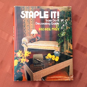 May include: A vintage book titled "STAPLE IT! Easy Do-It Decorating Guide" by Iris Ihde Frey. The cover features a colorful interior design scene with a bench, table, and plants. The book has 258 photographs, 79 drawings, and 18 color plates.