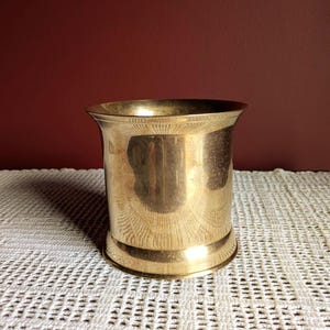 May include: A cylindrical brass container with a flared top and a rounded base. The container is highly reflective, with visible reflections of the surrounding environment. It sits on a white, textured surface against a dark red background.