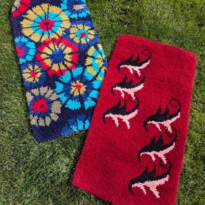 May include: Two rectangular, hand-tufted rugs. One is navy blue with a floral pattern in yellow, red, and blue. The other is red with a repeating black and pink stylized animal design. Both are on green grass.