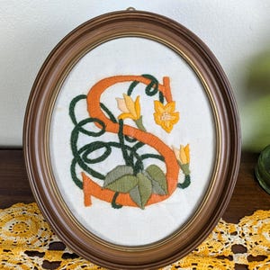 Letra inicial "S" naranja con ribete verde y marco ovalado, terminada en lana vintage.