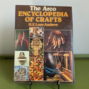 May include: A hardcover book titled "The Arco Encyclopedia of Crafts" with a brown cover and orange lettering. The cover features various craft examples, including weaving, mosaics, and beadwork. The book is displayed on a black metal stand.