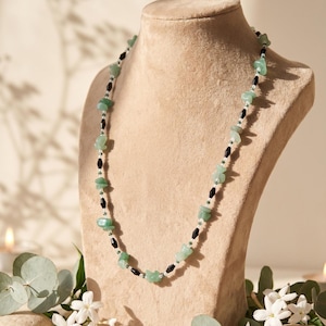 May include: A gemstone necklace displayed on a mannequin. The necklace features irregular, light green jade stones interspersed with black and white beads. The necklace is a delicate piece of jewelry.