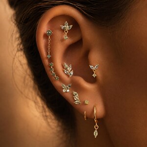 May include: A collection of gold-toned ear piercings featuring various designs including small dangling chains, leaf-shaped clusters, butterfly motifs, and marquise-cut green stones. The jewellery set includes multiple studs, hoops, and charms.