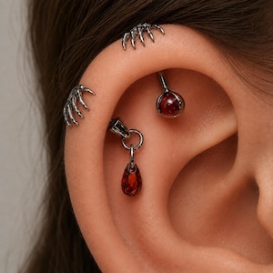 Op de afbeelding: Een set zilverkleurige oorpiercings met twee klauwachtige ontwerpen die het kraakbeen vastgrijpen, een bolvormige rode edelsteen in een klauwzetting en een bungelende druppelvormige rode edelsteen aan een kleine metalen ring.