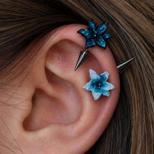 May include: Two ear piercings featuring flower-shaped studs with sharp, metallic spike backings. One flower is dark blue with glitter, and the other is a light blue and white gradient. The jewelry is positioned along the upper cartilage of the ear.