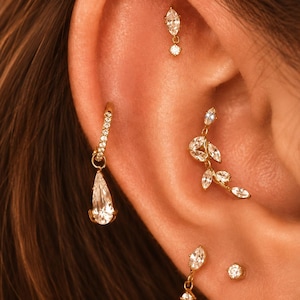 May include: A collection of gold-tone earrings featuring clear marquise and pear-shaped cubic zirconia stones. The set includes a hoop with a dangling pear-shaped stone, a leaf-shaped climber, a small stud with a dangling stone, and two round studs.