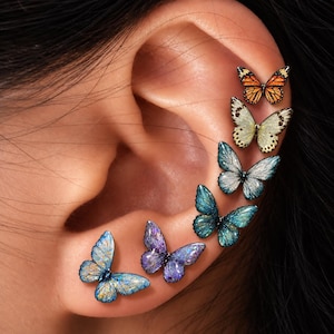 Könnte beinhalten: Ein Set aus sechs schmetterlingsförmigen Ohrsteckern, die entlang des äußeren Ohrrandes angeordnet sind. Die Schmetterlinge haben lebendige, glitzernde Oberflächen in Orange, Creme, Hellblau, Dunkelblau, Violett und Türkis mit schwarzen Mustern.