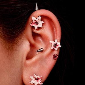 Op de afbeelding: Een collectie oorpiercings met drie witte bloemvormige knopjes met rode accenten, twee zilverkleurige metalen stekels en één kleine ronde rode edelsteenknop, gerangschikt langs het kraakbeen en de oorlel van het oor.