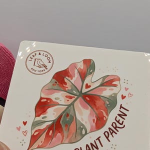 May include: A square coaster with a watercolor illustration of a leaf in shades of red, pink, and green, with small hearts around the leaf. The text "RARE PLANT PARENT" is written in a dark red script below the leaf. The coaster has the "LEAF & LOOM NEW YORK" logo.