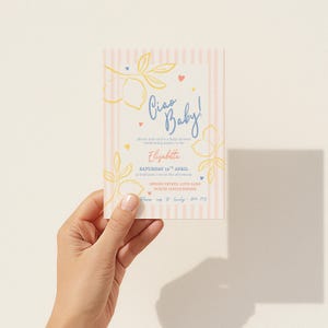 Puede incluir: Una invitaci&oacute;n para baby shower con un fondo de rayas verticales rosas y blancas. La invitaci&oacute;n presenta el texto "Ciao Baby!" en escritura azul, junto con ilustraciones florales y de limones. La invitaci&oacute;n se sostiene en una mano.