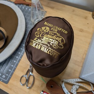 Puede incluir: Una caja de sombrero marr&oacute;n con letras doradas que dice "HAND CRAFTED FOR The Canadian Brim Co. Est. 2023 Bespoke Quality Headwear NOLAN WALCOTT." La caja est&aacute; sobre una superficie de madera con un sombrero, tijeras y una cinta m&eacute;trica.
