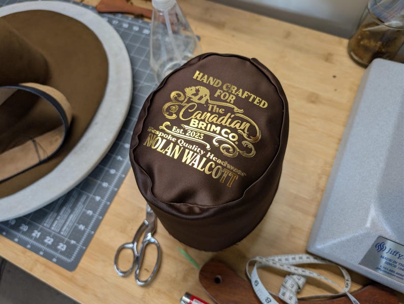 Puede incluir: Una caja de sombrero marr&oacute;n con letras doradas que dice "HAND CRAFTED FOR The Canadian Brim Co. Est. 2023 Bespoke Quality Headwear NOLAN WALCOTT." La caja est&aacute; sobre una superficie de madera con un sombrero, tijeras y una cinta m&eacute;trica.