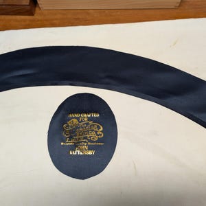Puede incluir: Una pieza de tela curva azul marino y un parche ovalado azul marino con texto dorado. El parche dice "HAND CRAFTED FOR The Canadian Brim Co. Bespoke Quality Headwear JOHN HATTERSBY."
