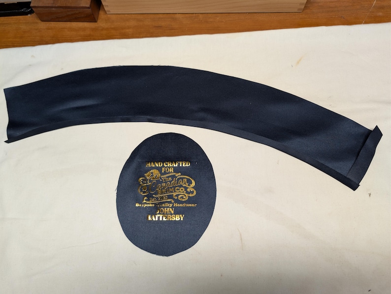 Puede incluir: Una pieza de tela curva azul marino y un parche ovalado azul marino con texto dorado. El parche dice "HAND CRAFTED FOR The Canadian Brim Co. Bespoke Quality Headwear JOHN HATTERSBY."
