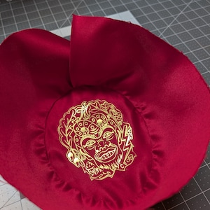Puede incluir: Un sombrero de tela rojo oscuro, similar al sat&eacute;n, con un gr&aacute;fico dorado de una cara con un rayo. El sombrero es circular con un ala ancha y una corona fruncida. El gr&aacute;fico est&aacute; centrado en la corona.