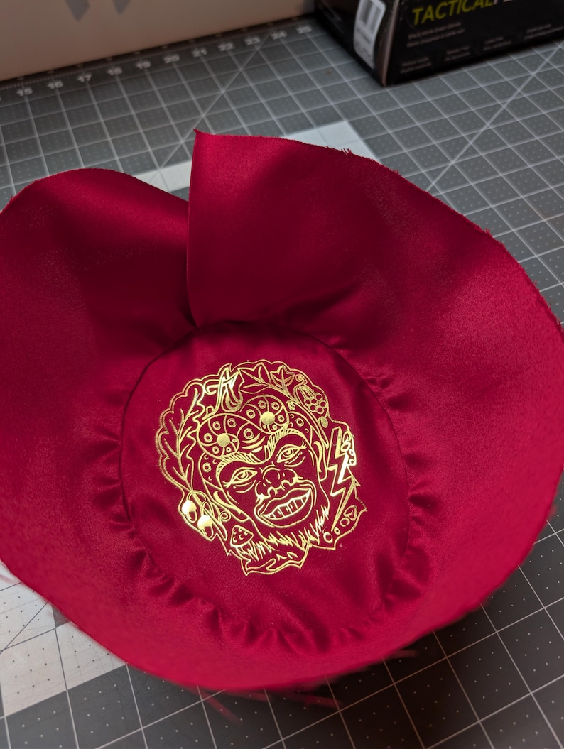 Puede incluir: Un sombrero de tela rojo oscuro, similar al sat&eacute;n, con un gr&aacute;fico dorado de una cara con un rayo. El sombrero es circular con un ala ancha y una corona fruncida. El gr&aacute;fico est&aacute; centrado en la corona.