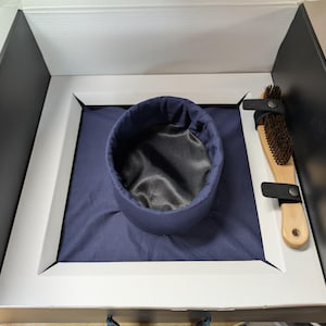 Puede incluir: Una caja abierta con un sombrero azul marino y un cepillo de madera. El sombrero tiene un interior de satén negro. El cepillo tiene cerdas oscuras y una correa de cuero. La caja tiene un marco blanco y un exterior negro.