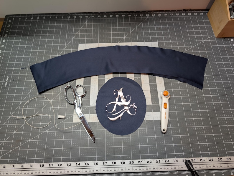 Puede incluir: Un trozo de tela azul marino, tijeras, un cortador rotatorio, un trozo de tela redondo con un dise&ntilde;o plateado, hilo y una barra de tiza blanca est&aacute;n dispuestos sobre una esterilla de corte gris. Los art&iacute;culos sugieren un proyecto de costura o manualidades.