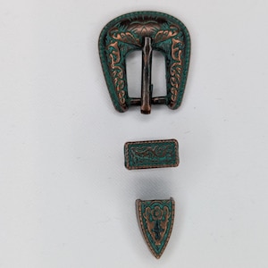 May include: A set of three decorative metal belt buckle components. The pieces have a copper and teal finish with ornate floral and scroll designs. The main buckle is arched, with a rectangular and a triangular piece below.