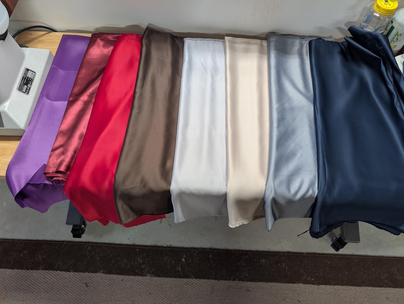 Puede incluir: Una colecci&oacute;n de telas plegadas en varios colores, incluyendo morado, burdeos, rojo, marr&oacute;n, blanco, beige, gris y azul marino. Las telas parecen ser de sat&eacute;n o un material liso similar, dispuestas sobre una mesa.