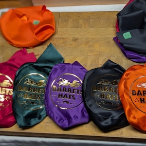 Puede incluir: Una variedad de bolsas para sombreros de colores, incluyendo naranja, negro, morado, verde y fucsia. Cada bolsa presenta el texto dorado "DARRETS HATS" dentro de un emblema circular. Las bolsas est&aacute;n dispuestas sobre una superficie de madera.