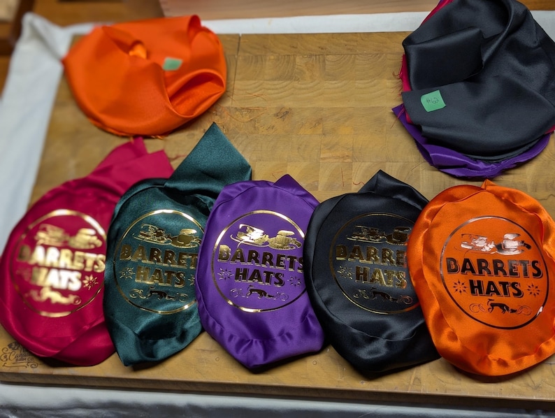 Puede incluir: Una variedad de bolsas para sombreros de colores, incluyendo naranja, negro, morado, verde y fucsia. Cada bolsa presenta el texto dorado "DARRETS HATS" dentro de un emblema circular. Las bolsas est&aacute;n dispuestas sobre una superficie de madera.