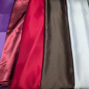 Puede incluir: Muestras de tela de sat&eacute;n de colores en tonos morado, burdeos, rojo, marr&oacute;n, azul claro y beige. La tela tiene una textura suave y brillante, y est&aacute; cuidadosamente dispuesta.