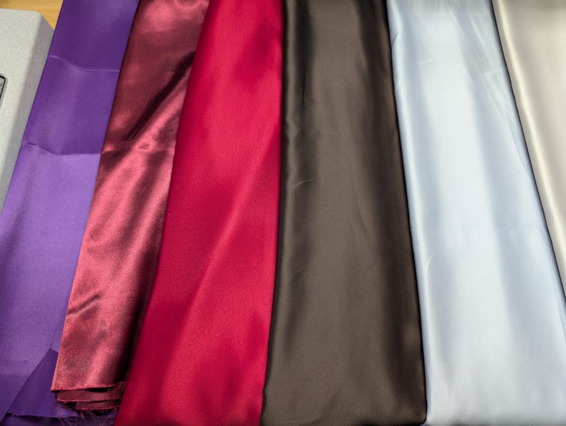 Puede incluir: Muestras de tela de sat&eacute;n de colores en tonos morado, burdeos, rojo, marr&oacute;n, azul claro y beige. La tela tiene una textura suave y brillante, y est&aacute; cuidadosamente dispuesta.