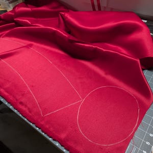 Puede incluir: Una vibrante tela de sat&eacute;n rojo con contornos de tiza para cortar. La tela est&aacute; drapeada sobre una esterilla de corte gris, con una caja negra y otras herramientas visibles en el fondo. Los contornos sugieren un patr&oacute;n para coser o crear.