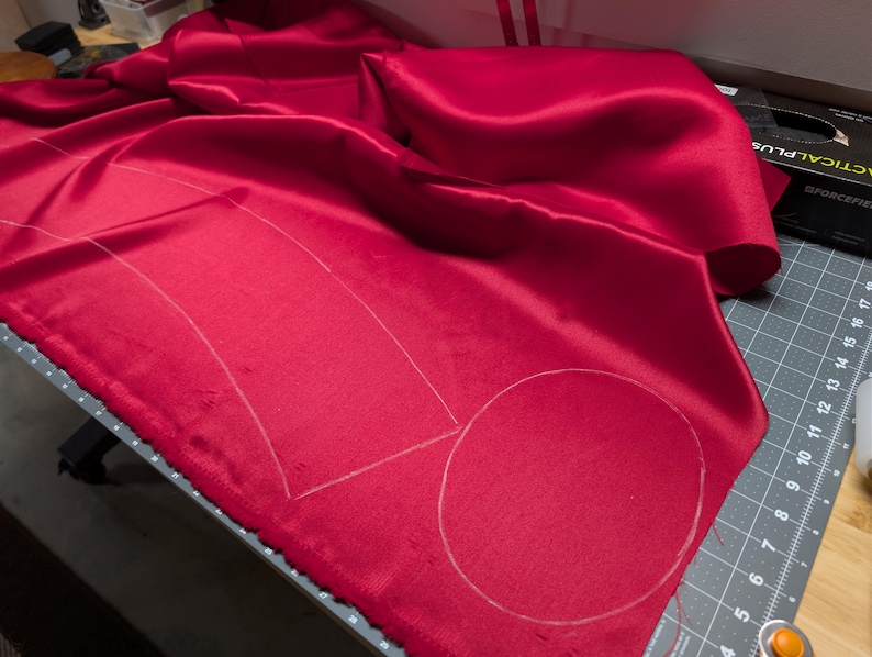 Puede incluir: Una vibrante tela de sat&eacute;n rojo con contornos de tiza para cortar. La tela est&aacute; drapeada sobre una esterilla de corte gris, con una caja negra y otras herramientas visibles en el fondo. Los contornos sugieren un patr&oacute;n para coser o crear.