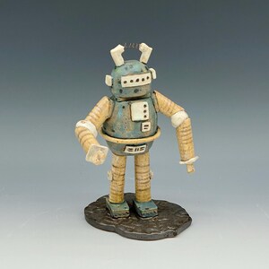 May include: A ceramic robot figurine with a speckled blue head and torso, tan segmented arms and legs, and white rectangular chest details. The robot features two antennae on top, a visor-like face, and stands on a dark brown textured base.