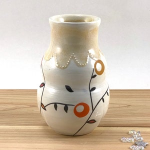 May include: A white ceramic vase with a cream colored rim and a scalloped edge. The vase is decorated with a floral design in black, orange, and red.