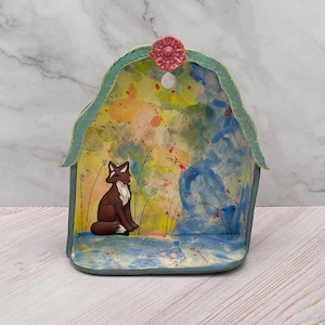 May include: A ceramic wall pocket featuring a hand-painted fox sitting in a field of tall grass. The background has a watercolor effect with yellow, blue, and green splashes. A small pink flower sits at the top center of the arched green frame.