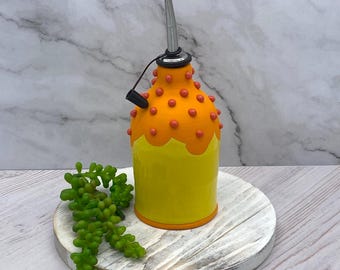 May include: A yellow ceramic oil bottle with a orange top and red dots. The bottle has a silver spout and is sitting on a white wooden round base.