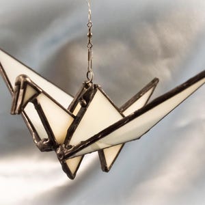 Pode incluir: Uma grulha de origami de vidro branco pendurada em uma corrente prateada. A grulha é feita de vidro e metal e tem um design delicado e intrincado.