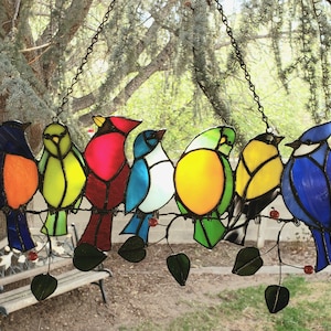 Könnte beinhalten: Ein Buntglas-Sonnenfänger mit sieben bunten Vögeln, die auf einem Ast mit grünen Blättern sitzen. Die Vögel sind rot, orange, gelb, grün, blau und lila.