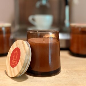 May include: A brown candle in a clear glass jar with a wooden lid. The lid has a red label with the text "Candles by Janice". The candle is next to a coffee machine and other candles.