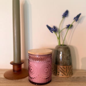 Può includere: Una candela rosa in un barattolo di vetro decorativo con coperchio in legno. Il barattolo ha un motivo in rilievo. Una candela verde e un portacandele marrone sono a sinistra. Un vaso con fiori blu è a destra.