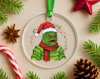Puede incluir: Adorno navideño redondo y transparente con una criatura verde con gorro de Papá Noel y bufanda roja. El adorno tiene una cinta verde para colgar. Decoración festiva con un tema de monstruo único.